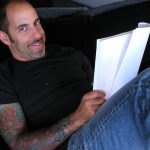 Man with short black hair and sleeve tattoo siting on a couch holding a script
