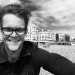 Black and white photo of a man with short hair and thick glasses on the beach